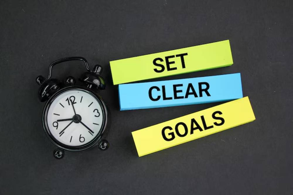 A black alarm clock next to colored sticky notes that read 'SET CLEAR GOALS' on a dark background.