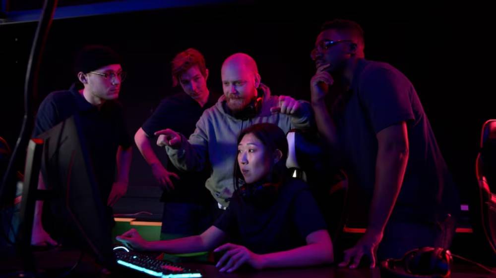 A group of gamers closely gathers around a computer, engaged in a discussion, with colorful lighting creating an energetic atmosphere.
