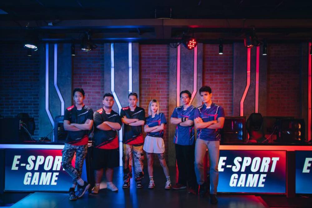 A group of six gamers poses confidently in an esports arena, with vibrant lighting and gaming equipment in the background.