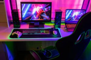 A vibrant gaming setup featuring a keyboard, mouse, Xbox controller, dual monitors, speakers, and an illuminated gaming chair.