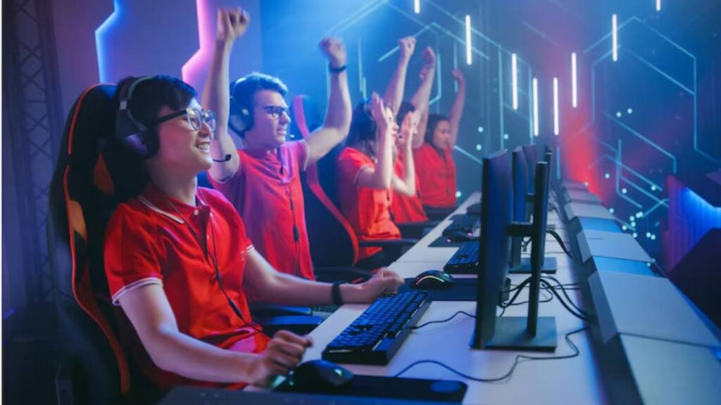 Four enthusiastic gamers in red shirts celebrate in a dimly lit gaming arena, surrounded by computers and colorful lights.