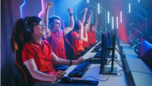 Four enthusiastic gamers in red shirts celebrate in a dimly lit gaming arena, surrounded by computers and colorful lights.