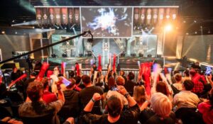 A lively esports event with an enthusiastic crowd cheering, holding foam sticks, and watching a large screen displaying game graphics.