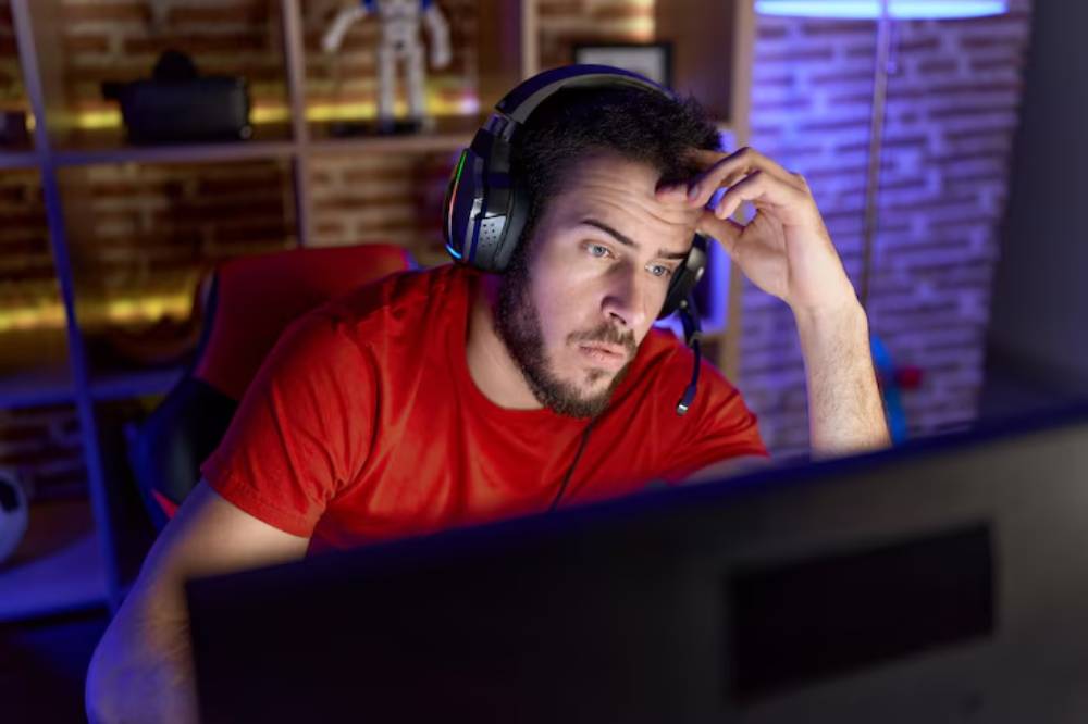 A person in a red shirt wearing headphones looking tensed, focused on a computer screen in a dimly lit, modern gaming setup.